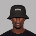 A person wearing a black bucket hat with a white rectangular patch that reads 'unus virtus' on a plain gray background.