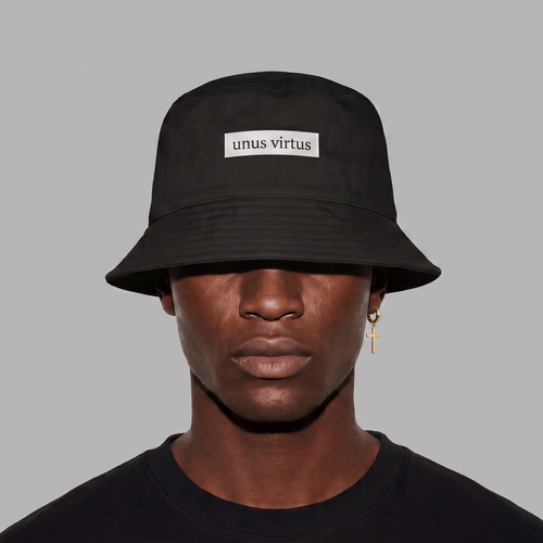 A person wearing a black bucket hat with a white rectangular patch that reads 'unus virtus' on a plain gray background.